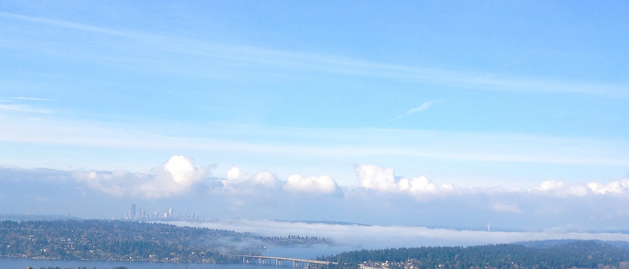 Daytime view of the Seattle skyline and Mercer Island across Lake Washington, with the I-90 floating bridge crossing the water in the foreground.